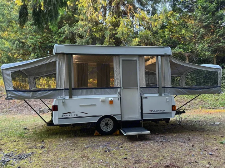 White Fleetwood Santa Fe pop-up RV rental set up in a forest. Ideal for camping adventures.. Fleetwood Santa Fe 2004