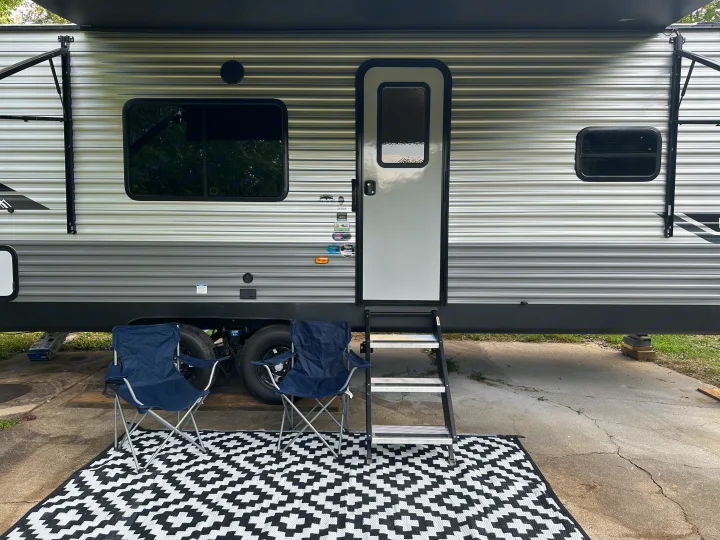 RV rental: The side of a travel trailer with an awning, steps, and an outdoor setup featuring a black/white mat and two blue chairs.. Coachmen 261BH 2024