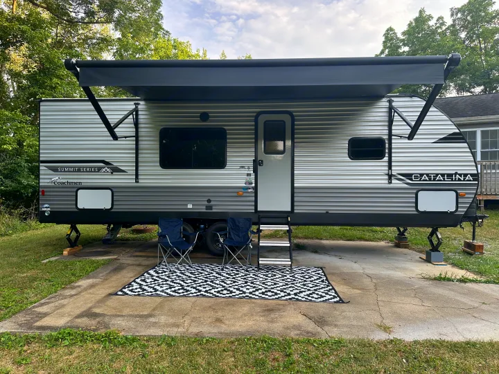 A comfortable RV rental setup: Coachmen Catalina travel trailer with extended awning, chairs, and outdoor rug on a concrete pad.. Coachmen 261BH 2024
