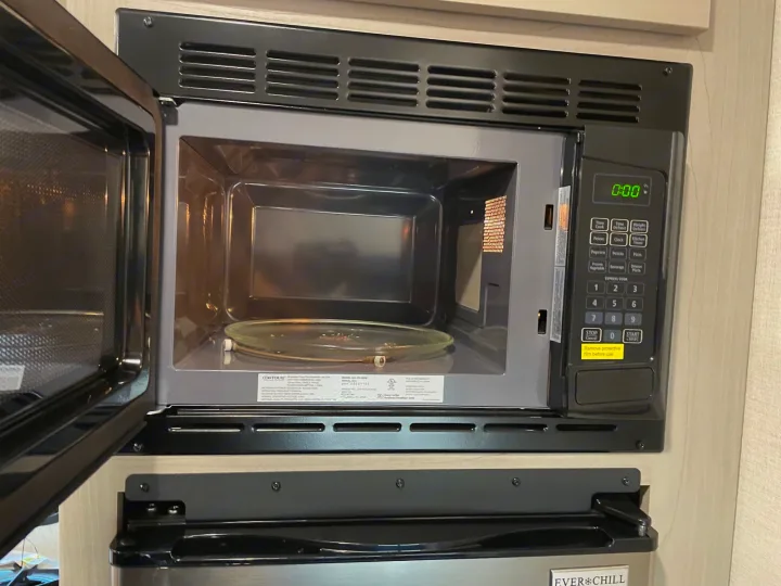 An open microwave oven, showing its glass turntable and 0:00 display, within an RV rental's compact kitchen.. Keystone Springdale 1800BH 2024