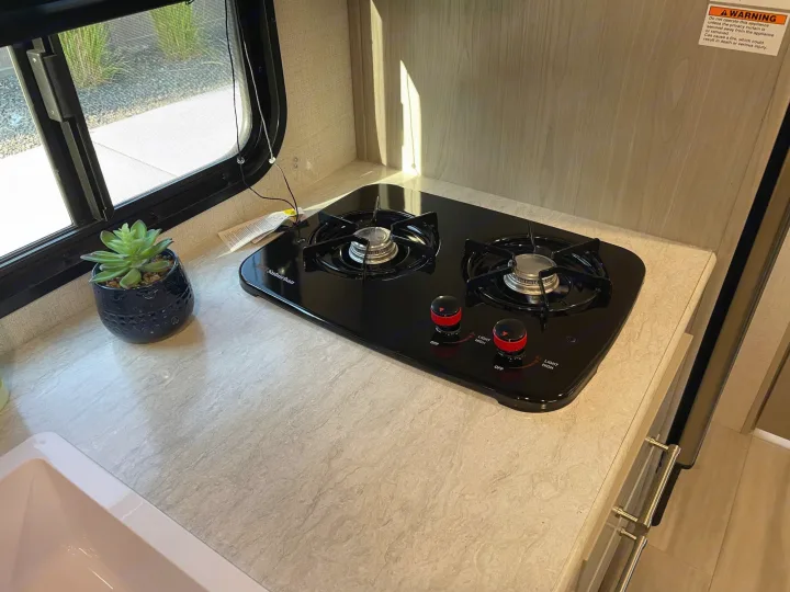 Compact kitchen in an RV rental, featuring a two-burner stovetop, light counter, and a window with a succulent plant.. Keystone Springdale 1800BH 2024