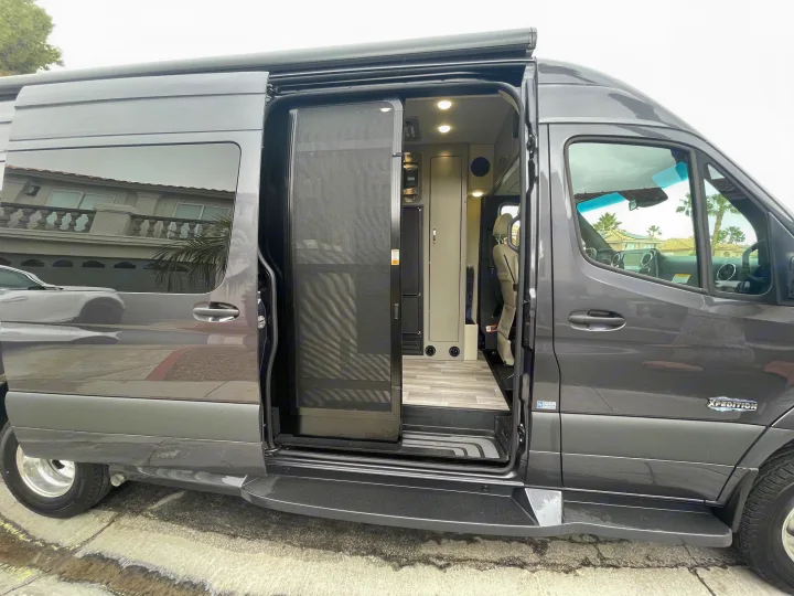 A modern gray rv rental camper van, with its side door open revealing the interior living space and a black screen door.. Holiday Rambler Expedition 2024