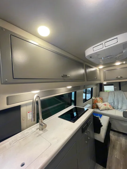 Bright RV rental interior: kitchen with sink, stovetop, and modern cabinets. Rear living area with sofa and AC unit visible.. Holiday Rambler Expedition 2024