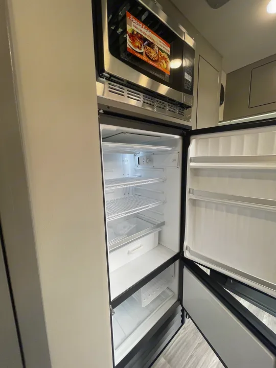 This RV rental kitchen features a microwave oven and an open, empty refrigerator with shelving and door bins.. Holiday Rambler Expedition 2024