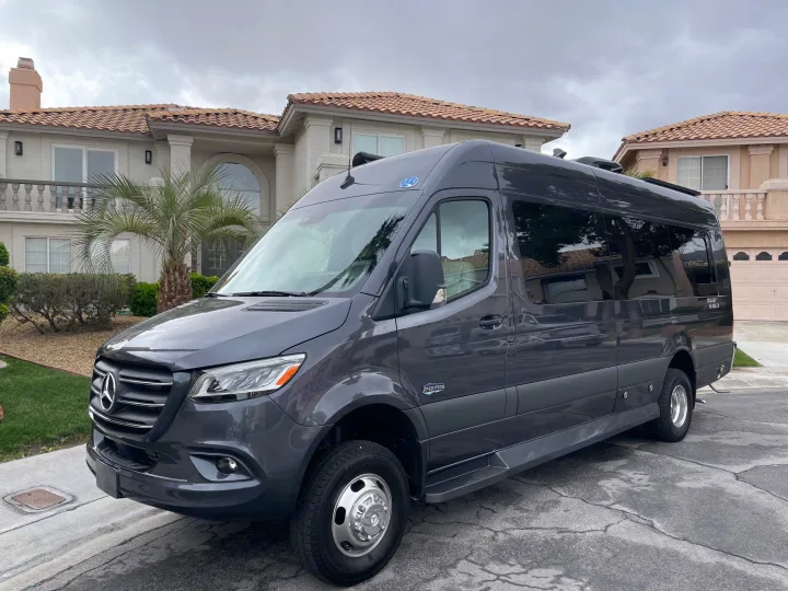 A sleek dark gray Mercedes-Benz Sprinter Expedition RV rental parked in front of a large suburban house on a cloudy day.. Holiday Rambler Expedition 2024