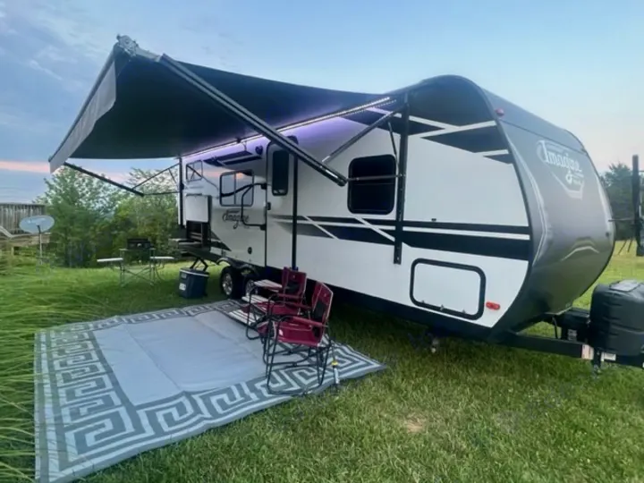This cozy rv rental setup features a white travel trailer with an extended awning, an outdoor rug, and chairs on a grassy site.. Grand Design Imagine XLS 2021
