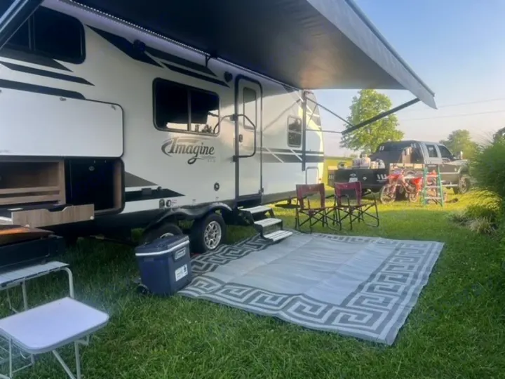 Imagine RV rental set up on grass. Travel trailer with extended awning, outdoor mat, cooler, chairs, and truck visible for camping.. Grand Design Imagine XLS 2021