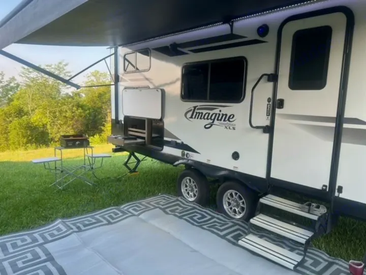RV rental setup with extended awning, outdoor kitchen, and a patterned mat on the grass.. Grand Design Imagine XLS 2021