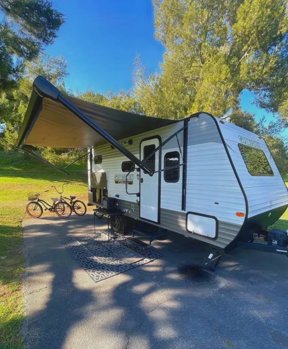 A white RV rental travel trailer with an extended black awning, two bikes, and an outdoor mat on asphalt under trees.. KZ SPORTSMEN CLASSIC 191BHK 2024