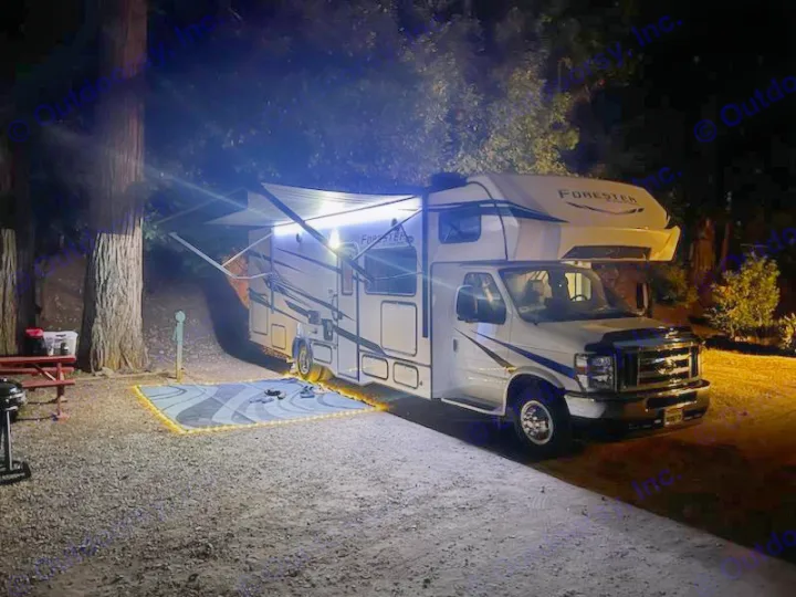 Nighttime at the campsite with an illuminated RV rental under its awning, complete with a cozy lit mat and picnic area.. Forester Classic 2023