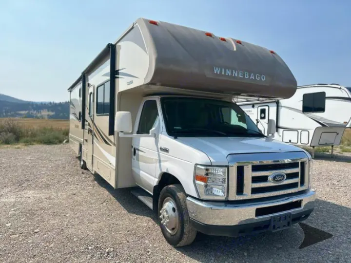 A white Winnebago Class C RV rental, ready for the open road. Parked on gravel with a natural landscape background.. Winnebago Minnie Winnie 2017