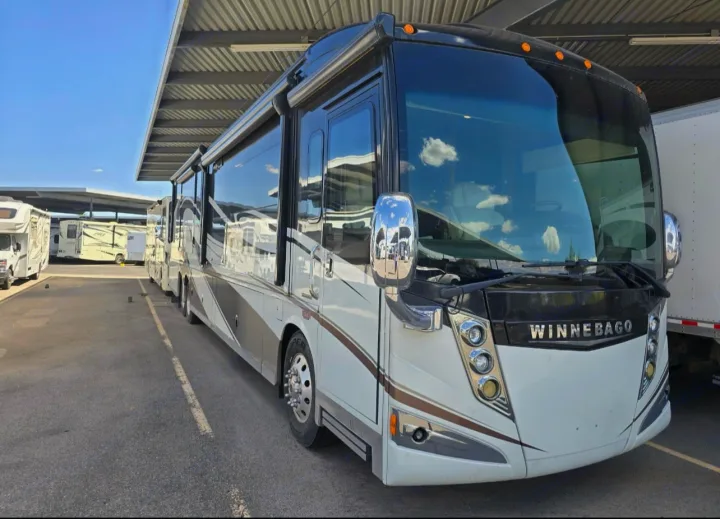 A large, luxurious Winnebago motorhome, white with brown accents, suitable for rv rental, parked under a shade structure.. Winnebago Ellipse 2016