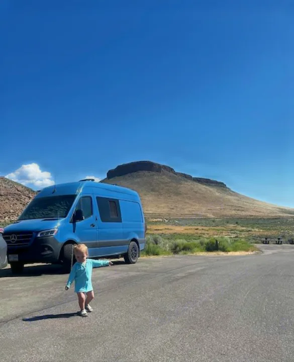 An RV rental, a vibrant blue van, is parked against a backdrop of a flat-topped mountain. A toddler walks in front.. Mercedes-Benz Sprinter 2021