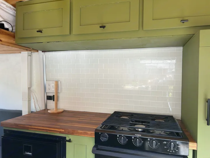 This modern RV rental kitchen features olive green cabinets, a wood countertop, white subway tile backsplash, and a Dometic stove/oven.. Ford Transit 2019