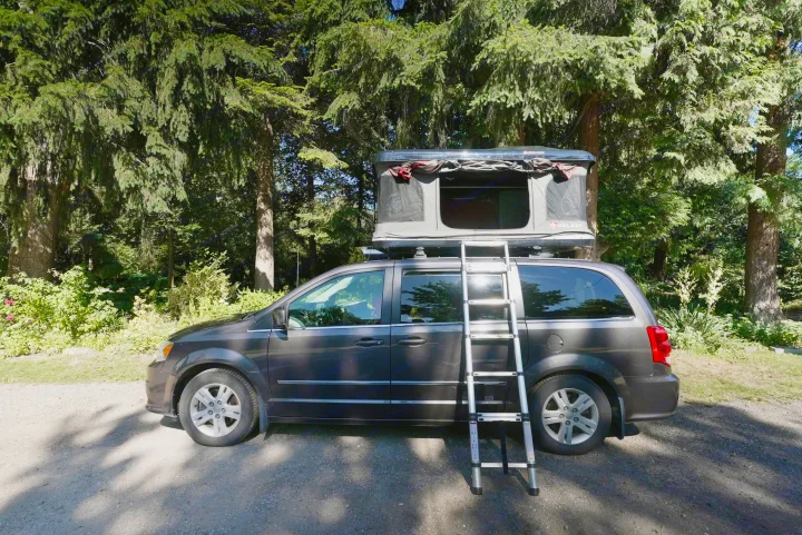 Minivan RV rental with a deployed rooftop tent and ladder, parked in a sunny, tree-filled camping spot.. Dodge Caravan 2015