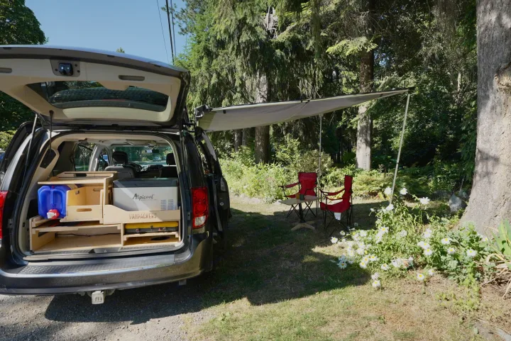 Van RV rental tailgate open, showcasing a custom "VANPACKERS" camping setup with cooler, water, stove, & awning shading chairs in nature.. Dodge Caravan 2015