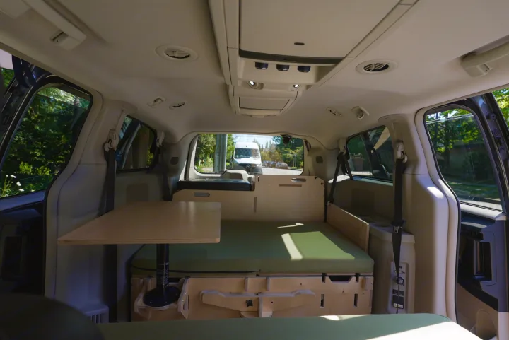 RV rental camper van interior featuring a wooden bed platform, green mattress, and collapsible table for compact travel.. Dodge Caravan 2015
