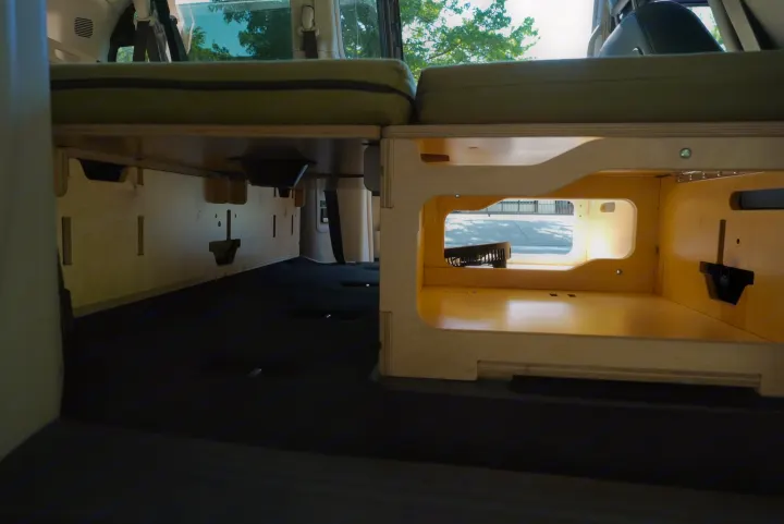 Inside an rv rental, a light wood bed frame supports green cushions, with visible modular under-bed storage compartments.. Dodge Caravan 2015