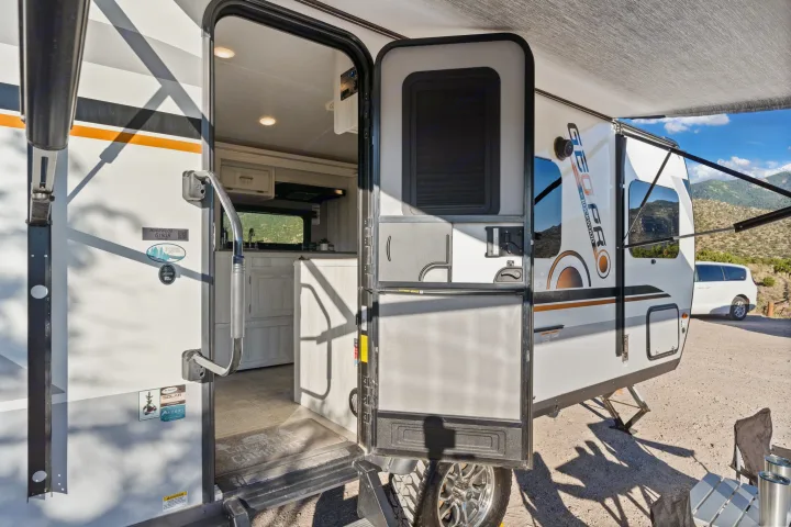 Open door of a modern GEO PRO Rockwood travel trailer RV rental, revealing the kitchen and a scenic mountain campsite setup.. Rockwood G19QB 2021