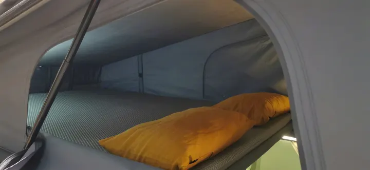 Experience comfort in this RV rental's pop-top sleeping area, featuring a mattress and two orange pillows, ready for rest.. Renault TRAFIC 2023