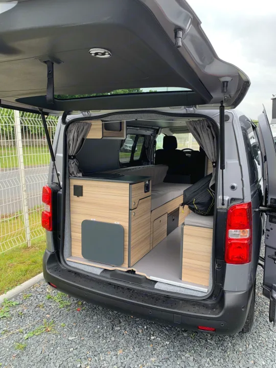 Inside this modern RV rental: a grey camper van with its tailgate open, showcasing a modular wood-grain conversion, bed, and storage.. opel L'OTAGO (Caen) 2024