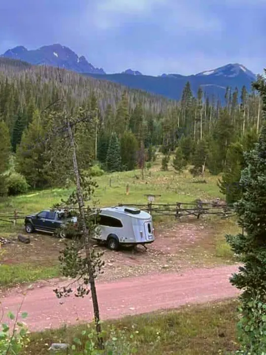 A grey teardrop RV rental and pickup truck are parked on a dirt road, surrounded by a mountain forest. Perfect for outdoor travel.. Airstream Basecamp 2023