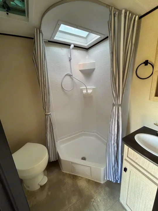 Compact RV rental bathroom featuring a shower with a skylight, toilet, and sink, accented by a striped curtain.. Coachmen Apex Nano 2023