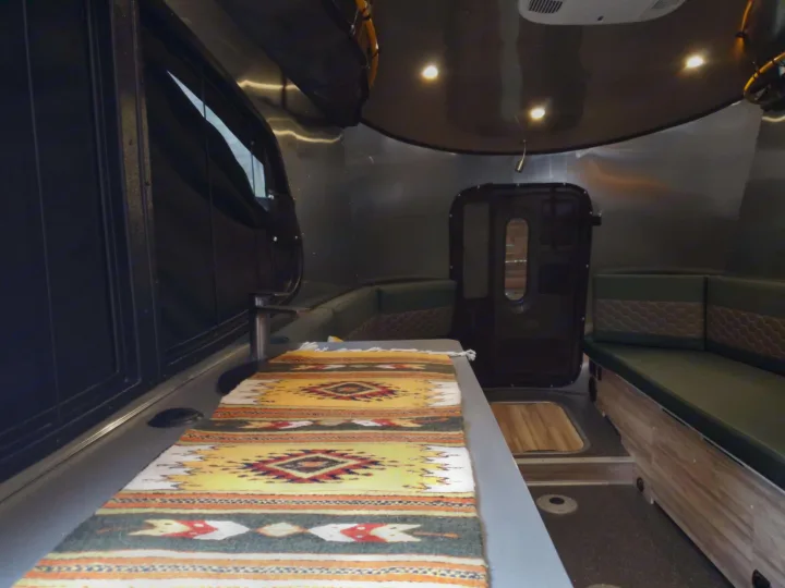 Explore this inviting RV rental interior! Features a table with a vibrant rug, comfy green seating, and ample space.. Airstream Basecamp 2023