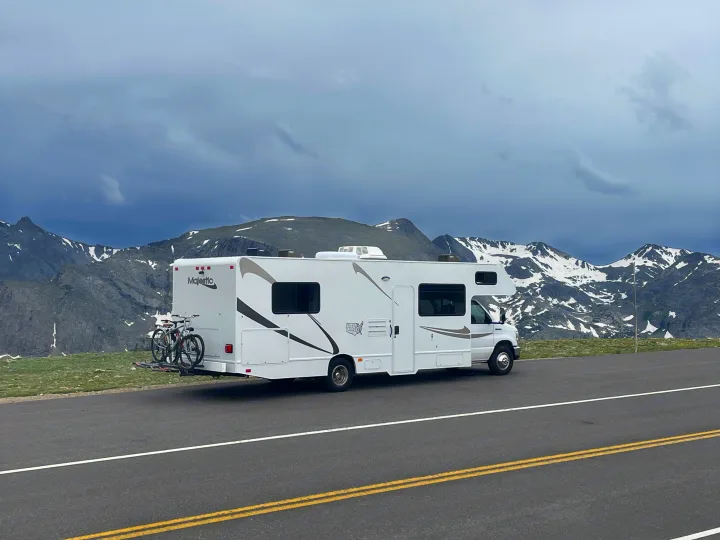 This RV rental is ready for mountain adventures! White RV with bikes parked on a scenic road with snowy peaks.. Thor Majestic 2019