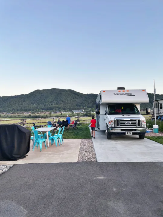 Family enjoying their Majestic RV rental at a mountain campsite. Features a patio with turquoise chairs and a grill cover.. Thor Majestic 2019