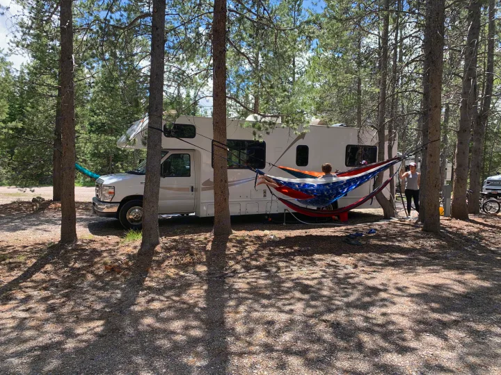 RV rental parked in a wooded campsite with colorful hammocks, including a child relaxing and a woman standing nearby.. Thor Majestic 2019