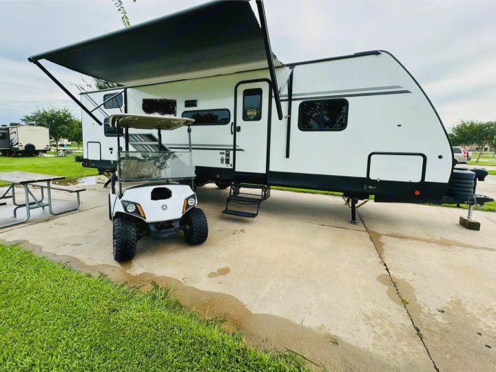 RV rental ready for adventure: A white travel trailer with awning deployed, parked next to a golf cart on a concrete campsite.. Dutchmen Kodiak SE 2024