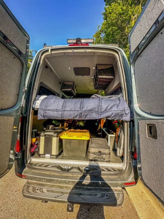 RV rental: Back of a cozy camper van showing a raised bed, under-bed storage, and custom interior for road trip adventures.. Mercedes-Benz Sprinter 2024