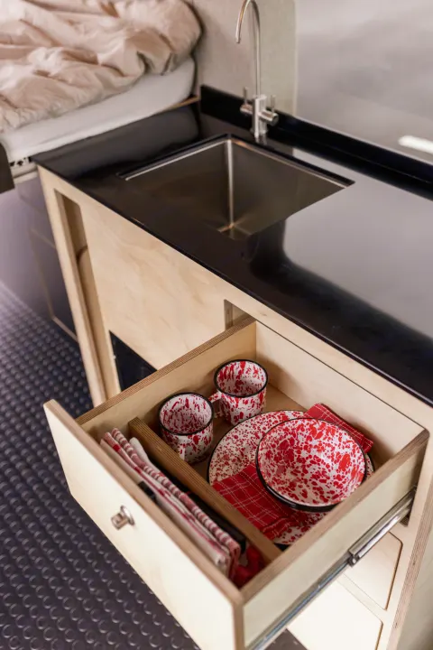 Compact RV rental kitchen features a sink and a drawer with red speckled enamelware dishes & plaid napkins.. Ford Transit 2020