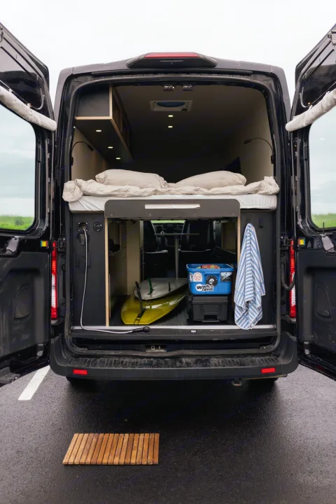 RV rental van's open rear reveals bed, gear storage including surfboards, and an outdoor shower.. Ford Transit 2020