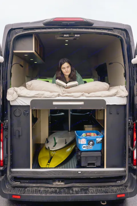 Enjoying an RV rental! A person reads on a cozy bed in this customized camper van, with surfboards & storage below.. Ford Transit 2020