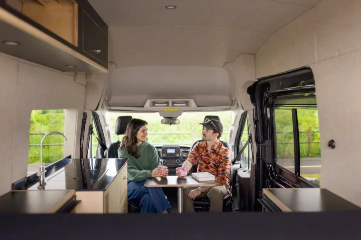 Enjoying an RV rental: A couple relaxes in their van's modern interior with a kitchen and dining table, surrounded by nature.. Ford Transit 2020