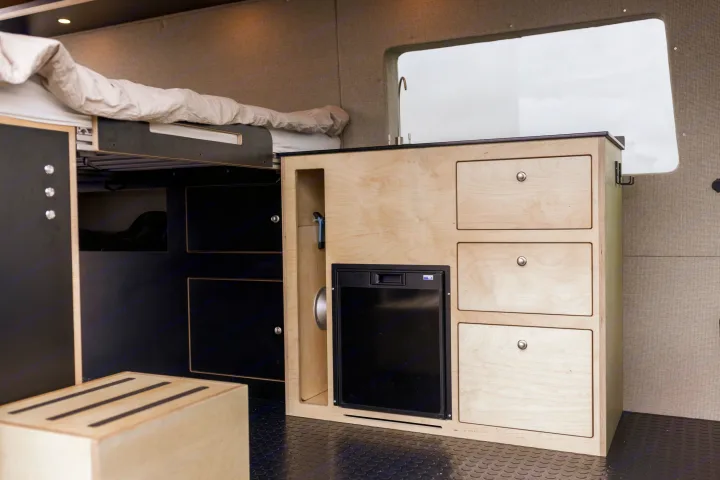 Compact RV rental interior with a bed, light wood kitchen featuring a fridge & sink, and ample storage solutions.. Ford Transit 2020