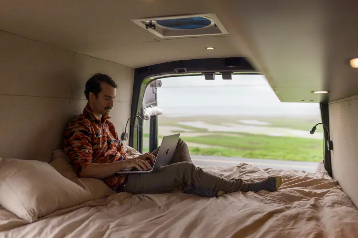 A man on a comfortable bed inside an RV, focused on his laptop. The rear opens to a scenic outdoor landscape. Ideal for RV rental.. Ford Transit 2020