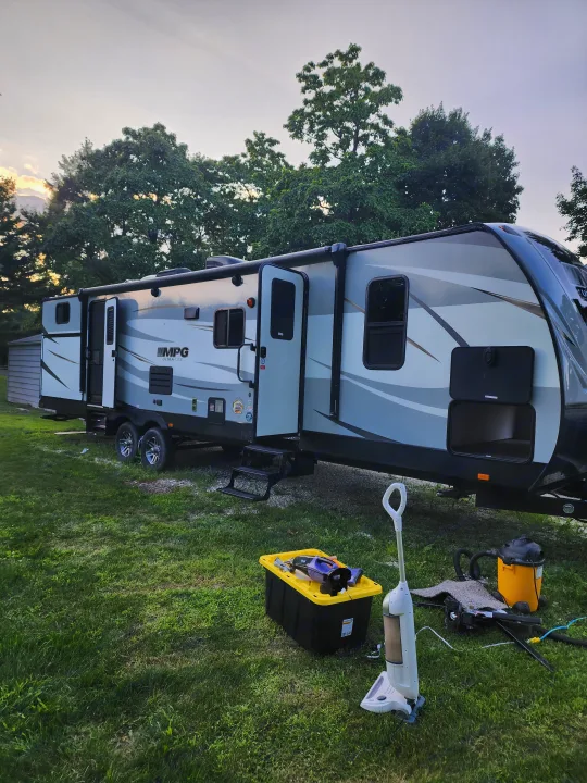 Grey and white travel trailer RV rental parked on grass. Its door is open, and cleaning equipment is visible nearby.. Cruiser RV MPG Ultra Lite 2020