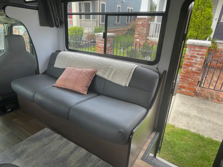 Cozy RV rental interior: gray sofa, pink pillow, blanket, and a large window with a view of a neighborhood street.. Thor Majestic 2019