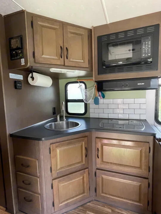 Modern rv rental kitchen featuring a corner layout with sink, microwave, stovetop, and light wood cabinets with subway tile backsplash.. Thor Majestic 2019