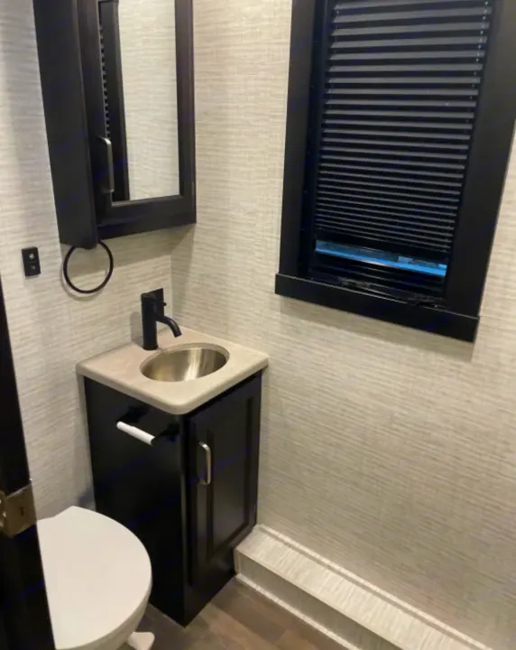 Compact RV rental bathroom featuring a dark vanity with sink, mirror, toilet, and window with blinds.. Entegra Coach Other 2024