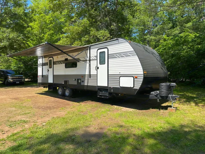 Spacious travel trailer RV rental with awning extended, parked on a dirt and grass lot amidst green trees.. Aspen Trail LE 2022