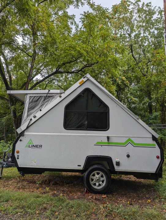 A gray A-liner A-frame pop-up camper, suitable for rv rental, parked in a lush, tree-filled setting with rain droplets visible.. A Liner A Liner Popup Trailer 2022