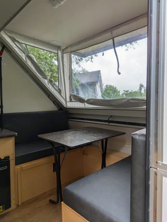 Inside an RV rental: a compact, light-filled dinette with black bench seating and a patterned table, looking out to trees.. A Liner A Liner Popup Trailer 2022