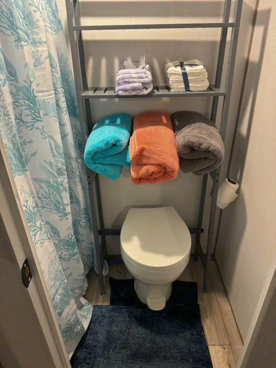 RV rental bathroom: compact toilet, storage shelf with neatly folded towels, and a blue coral shower curtain.. Dutchmen Coleman 17B 2024