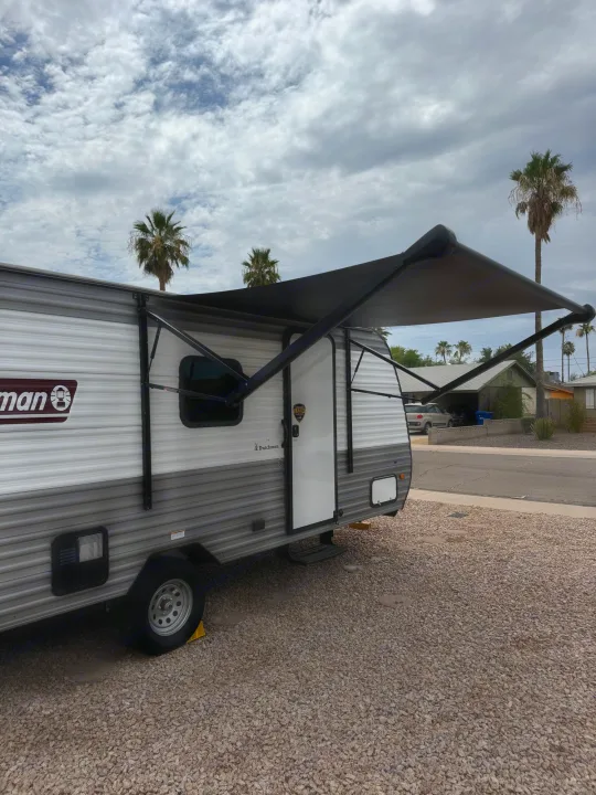 A gray and white Coleman travel trailer with its awning extended, parked on gravel. Consider this RV rental for your next trip.. Dutchmen Coleman 17B 2024