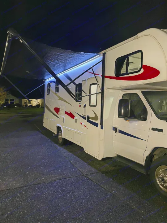 An RV rental, a white Class C motorhome, parked at night with its extended awning and bright blue LED lights.. Forester Forester Motorhome 2019