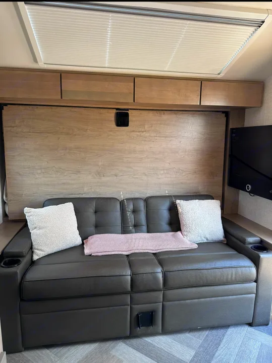 Inside this RV rental: a comfy dark sofa with pillows and blanket, smart overhead storage, and a mounted TV. Note the murphy bed wall.. Ember Overland 2023